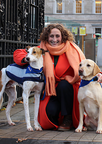 Person in an orange coat and scarf kneeling beside two dogs wearing blue vests, one with a red backpack, in front of a black iron fence and building with large windows—suggesting a service or therapy dog setting in an urban environment.