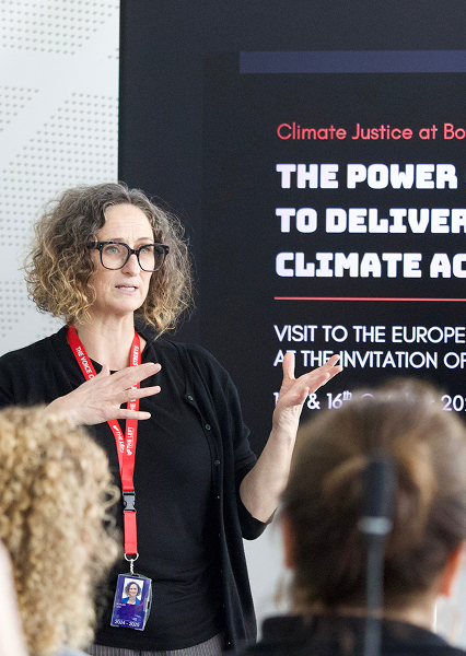 Speaker with red lanyard presenting on climate justice and action, with audience members visible and a screen displaying event details behind them.