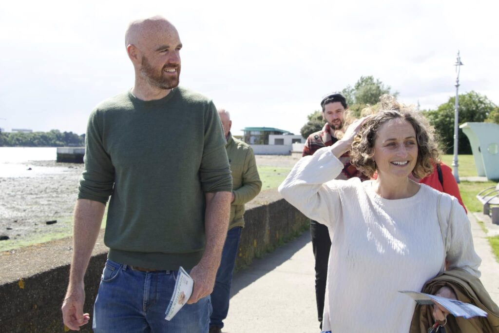 Group of people walking along a waterfront path; one holds a brochure, another a piece of paper, with grassy areas, a rocky shore, and a building in the background, suggesting a casual outdoor gathering or tour.