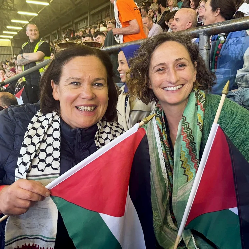 Two people holding Palestinian flags at a public gathering, wearing traditional-patterned scarves, with a crowd seated in a stadium behind them.