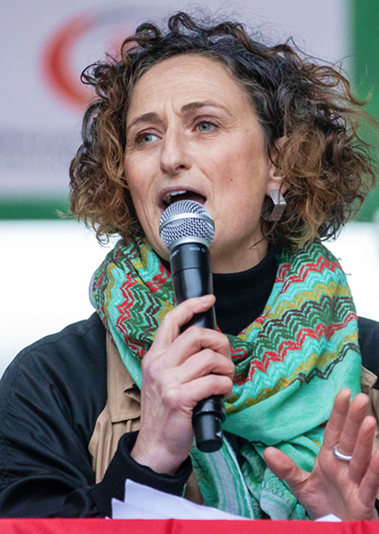 Person in a beige jacket and colorful zigzag scarf speaking into a microphone, with a blurred red and green logo in the background.
