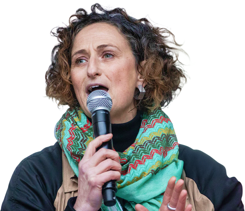 Person in a beige jacket and colorful zigzag scarf speaking into a microphone, captured mid-presentation.