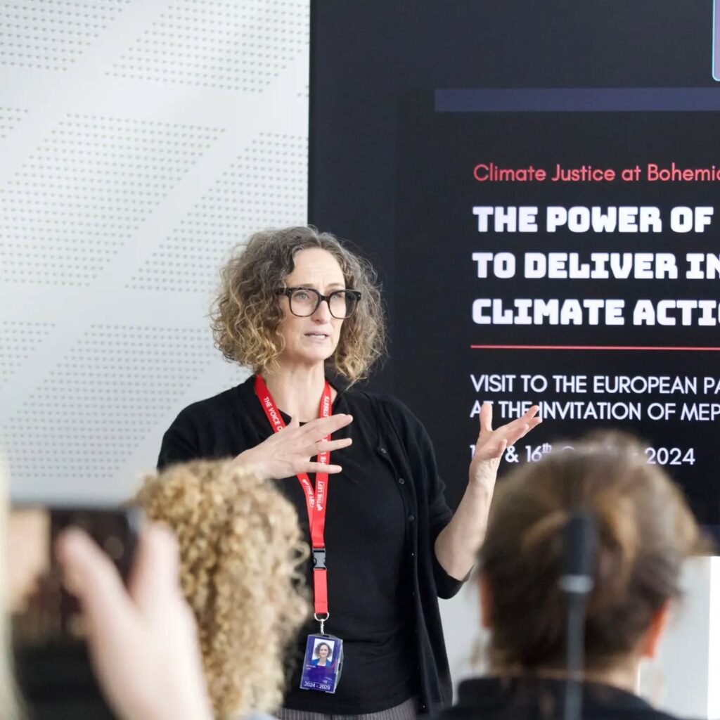 Speaker with red lanyard presenting on climate justice and action, with audience members visible and a screen displaying event details behind them.