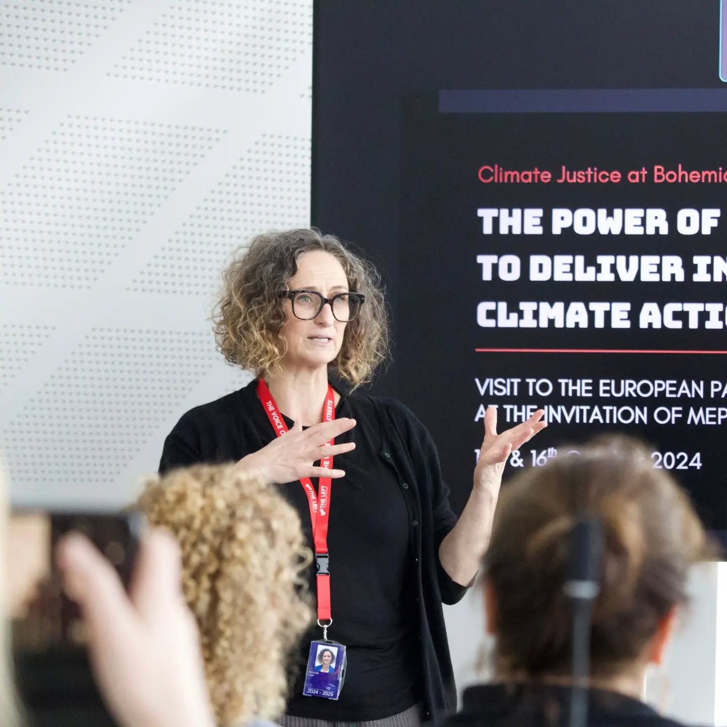 Speaker with red lanyard presenting on climate justice and action, with audience members visible and a screen displaying event details behind them.