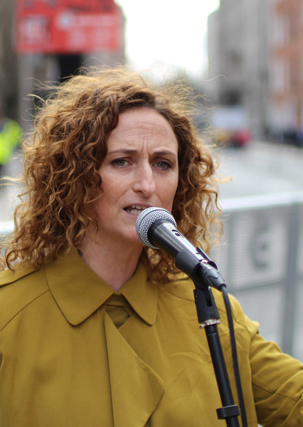 Person with curly hair in a mustard coat speaking into a microphone in an urban outdoor setting, with buildings and blurred figures in the background.