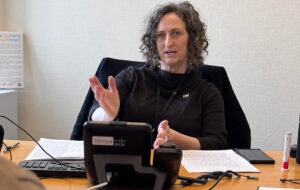 Person seated at a desk gesturing during a discussion, surrounded by office supplies including a Cisco phone, papers, and a keyboard.