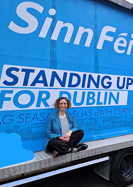 Person in a blue jacket and black pants sits cross-legged on a platform in front of a large blue Sinn Féin banner that reads “STANDING UP FOR DUBLIN” and its Irish translation “AG SEASAMH SUAS d'ÁTH CLIATH,” suggesting a political campaign or event.