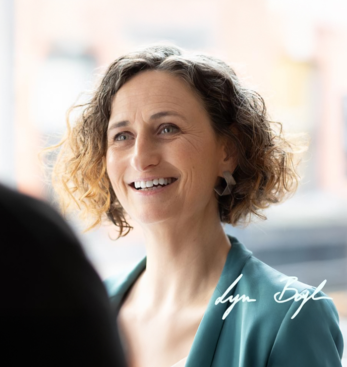 Person with curly hair in a teal blazer and earrings, indoors with soft natural light and a signature reading “Lyn Boyl” in the corner.