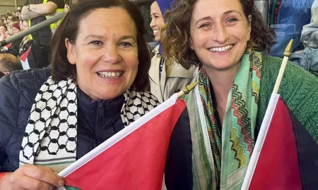 Two people holding Palestinian flags at a public gathering, wearing traditional-patterned scarves, with a crowd seated in a stadium behind them.