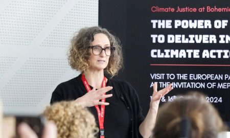 Speaker with red lanyard presenting on climate justice and action, with audience members visible and a screen displaying event details behind them.