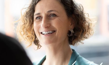 Person with curly hair in a teal blazer and earrings, indoors with soft natural light and a signature reading “Lyn Boyl” in the corner.